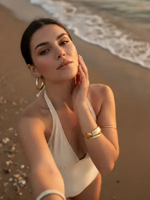 A woman stands gracefully on the sandy beach by the water, capturing a lifestyle beauty moment with her smartphone. The camera looks down on her, giving a high perspective typical of a handheld selfie. She tilts her head slightly upwards, gazing at the camera, while her hand delicately brushes the side of her face, fingers tracing the curve of her cheek and jawline. Her shoulders are relaxed, creating an elegant pose in the frame that captures her from the chest up.

Dressed in a cream halter neck swimsuit or summer top, her open shoulders and exposed collarbones exude a sense of summer chic. Adorning her look are large gold hoop earrings, a thick sculptural gold bracelet, a light marble-style bangle, and a delicate metallic arm bracelet above her elbow. Her makeup is soft and glamorous, perfect for a beach setting: her skin glows with a warm sun-kissed radiance, peach blush accentuates her cheekbones, clean sculpted brows frame her eyes, subtle winged eyeliner enhances her gaze, long lashes flutter, and her lips shimmer with satin finish.

In the background, the wet beach sand offers natural textures, with a thin line of sea foam marking the approach of a small wave behind her. Small shells are scattered on the sand, adding to the coastal atmosphere. The lighting captures the magic of a sunset or golden hour, casting warm highlights on her skin and creating soft shadows that play around her facial features. The shallow depth of field adds to the candid summer vacation mood, blending a realistic DSLR smartphone hybrid aesthetic seamlessly. 