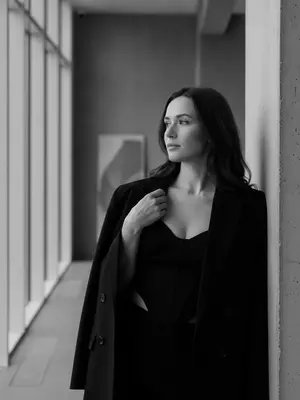 Женщина стоит в углу комнаты, наклонившись к стене. Ее лицо, с выражением спокойной уверенности, обращено к окну, вдалеке. На ней большой черный пиджак, небрежно надетый на плечи, и топ-корсет с V-образным вырезом. Рука лежит на пиджаке, поддерживая ее. Комната пронизана естественным светом из больших окон слева. Общая атмосфера внутри - стильная и спокойная, подчеркивающая элегантность и минимализм. Женщина позиционируется справа от центра кадра. Освещение мягкое, придающее ощущение простоты и минимализма. Черно-белая палитра фотографии выделяет контрасты и текстуры, создавая загадочную, но элегантную атмосферу. Портрет выполнен в ультрареалистичном 8K разрешении. 