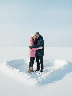 На фоне светло-голубого зимнего неба без облачка лежит плотный белоснежный снег, вырезанный в форме сердца. В середине этого сердца стоит пара, обнимаясь и улыбаясь в объектив камеры. Их стильная зимняя одежда выглядит современно, подчеркивая их хорошо видимые лица, на которых играют радостные улыбки.

Дневной свет придает снегу и тканям одежды мягкие холодные оттенки, подчеркивая каждую деталь. Атмосфера на фотографии наполнена уютом, любовью и спонтанностью зимнего момента. Снимок выполнен в реалистичном стиле, с фокусом на людях, создавая ощущение живого кадра, словно снятого с экшн-камеры или смартфона. Вертикальный формат добавляет динамики сцене.

Женщина одета в розовый пуховик с белой шапкой, украшенной помпоном, в то время как мужчина выбрал темный пуховик и шапку темного оттенка. 