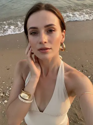 A woman stands gracefully on the sandy beach by the water, capturing a lifestyle beauty moment with her smartphone. The camera looks down on her, giving a high perspective typical of a handheld selfie. She tilts her head slightly upwards, gazing at the camera, while her hand delicately brushes the side of her face, fingers tracing the curve of her cheek and jawline. Her shoulders are relaxed, creating an elegant pose in the frame that captures her from the chest up.

Dressed in a cream halter neck swimsuit or summer top, her open shoulders and exposed collarbones exude a sense of summer chic. Adorning her look are large gold hoop earrings, a thick sculptural gold bracelet, a light marble-style bangle, and a delicate metallic arm bracelet above her elbow. Her makeup is soft and glamorous, perfect for a beach setting: her skin glows with a warm sun-kissed radiance, peach blush accentuates her cheekbones, clean sculpted brows frame her eyes, subtle winged eyeliner enhances her gaze, long lashes flutter, and her lips shimmer with satin finish.

In the background, the wet beach sand offers natural textures, with a thin line of sea foam marking the approach of a small wave behind her. Small shells are scattered on the sand, adding to the coastal atmosphere. The lighting captures the magic of a sunset or golden hour, casting warm highlights on her skin and creating soft shadows that play around her facial features. The shallow depth of field adds to the candid summer vacation mood, blending a realistic DSLR smartphone hybrid aesthetic seamlessly. 