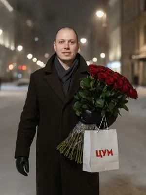 Мужчина в темно-коричневом пальто, шагает по улице, покрытой снегом, под ночным небом. В его руках громадный букет красных роз с зелеными листьями и пакет отмеченный логотипом "ЦУМ". Его взгляд сосредоточен, излучая уверенность и спокойствие, с легкой улыбкой на лице. Вспышка освещает его лицо и аксессуары, создавая игру света и тени, подчеркивая контуры лица и верхней одежды. На заднем плане пространство города, покрытое снегом, с приглушенными огнями уличных фонарей и пустыми улицами, что придает ощущение уединения и стиля. Съемка выполнена на объективе 85 мм с диафрагмой f/1.4, с акцентом на лице и аксессуарах мужчины. 