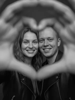 A candid and joyful couple stands out in a dreamy and intimate setting, exuding a romantic vibe that feels straight out of a movie scene. Their genuine smiles radiate warmth as they gaze directly at the viewer, creating a sense of connection. In the forefront of the image, the couple's intertwined hands subtly blur into a heart shape, delicately framing their beaming faces, adding a touch of sweetness to the overall composition. 