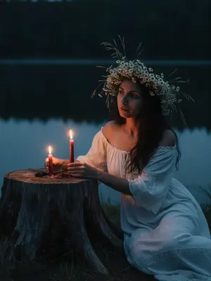 In the tranquil twilight by a serene lake, a woman is seated next to an ancient tree stump. Her posture exudes a dreamy relaxation, leaning slightly forward, one arm casually draped over her knee, the other hand delicately cradling a slender crimson candle beside another flickering flame on the stump. She wears a vintage off-shoulder peasant dress in white, adorned with intricate lace accents and gentle fabric drapes. Her hair is styled with a natural softness, featuring loose strands framing her face.

Adorning her head is a lavish crown of wildflowers, crafted from chamomile blooms, meadow grass, and delicate white blossoms. The background is enveloped by a dark forest and the placid waters of the lake, while the warm glow of the candles casts a gentle light on her face and hands, creating deep blue shadows in the mystical ambiance of a summer night.

Captured with the night mode of an iPhone, the image boasts a realistic grain in low light, with adjusted exposure to maintain sharpness and clarity, devoid of flash, blur, or bokeh effects. 