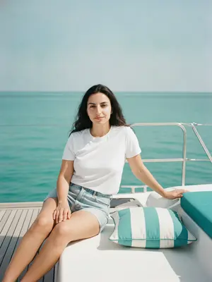 На палубе катамарана модель сидит удобно, одна рука отдыхает на белом борту, а другая лежит на ноге. Ее корпус немного повернут к камере, глаза смотрят прямо в объектив, лицо украшает спокойная улыбка. Она одета в белую футболку и шорты.

Камера находится на уровне груди или немного ниже, а в кадре видно большую часть тела модели и окружающей среды. Фон занимает большую часть кадра, с небом в верхней части. Горизонтальная линия делит кадр пополам. Справа от модели виден кусочек катамарана с металлическими опорами и бирюзовым сиденьем, а на палубе заметна подушка с бирюзово-белыми полосами.

Используется стандартный объектив с фокусным расстоянием около 35–50 мм и диафрагмой примерно f/4–f/5.6. Глубина резкости умеренная: модель четко в фокусе, а фон (море и небо) слегка размыт, без явного боке. 