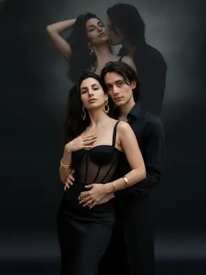 На фотографии запечатлены мужчина и женщина, стоящие в элегантной позе. Женщина одета в корсет с прозрачными вставками и длинную черную юбку, облегающую ее фигуру. Мужчина, находясь немного позади, обнимает ее за талию и устремленно смотрит в объектив камеры. Ее рука легко касается области груди, в то время как другая легко лежит на бедрах, создавая динамичную позу, выражающую уверенность и привлекательность.

Женщина украшена массивными золотыми серьгами, а на ее руках блестят золотые браслеты и кольца. Мужчина одет в черную рубашку и свободные брюки, прикрывая меньшую часть фона. Фон изображения темный и минималистичный, с мягкой и тусклой проекцией той же пары в верхней части, но в другой позе.

Естественное освещение выделяет мужчину и женщину на верхней части изображения, они занимают значительное пространство. Женщина соблазнительно смотрит, держа одной рукой волосы, а мужчина, целуя ее в шею, добавляет интимности этой сцене. Детали изображения максимально проработаны, придавая ему реалистичность. 