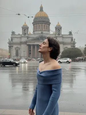 Фотография на улице Санкт-Петербурга после дождя, передающая атмосферу города. На снимке запечатлен человек в стильной зоне. Он стоит у фонарного столба, обрамленного сырым камнем, который отражает отблески влажного асфальта. Человек одет в черное пальто и широкополую шляпу, ладонью касается края головного убора. Его взгляд устремлен вдаль, словно он ожидает кого-то или что-то. Волосы чуть взмокли от оставшихся после дождя капель. На лице бледный свет от фонаря, создающий загадочную атмосферу. Вокруг витает запах свежести, а на фоне зданий виднеется туман, придающий картины загадочности и некой мистической притягательности. 
