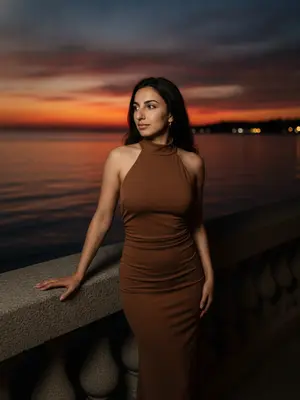 На набережной, где тихо вечерело, фото сделано с высоты, показывая женщину в элегантном обрамлении заката. Ее голова повернута, взгляд уходит вдаль, а улыбка легка. В макияже густые ресницы и блеск на губах. 
Фото вертикальное, охватывает женщину от бедер до головы. Она стоит вполоборота, одной рукой касается каменного парапета. За ее спиной широкая водная гладь, отражающая закатное небо и темные силуэты береговой линии. 
Небо и вода играют главные роли - от синего вверху до огненного оранжевого на горизонте. Свет приглушенный, идущий от заката, создает мягкую подсветку и теплый контур на волосах. 
Одежда и лицо освещены рассеянным светом, кожа выглядит матовой. Каменный парапет и берег в тени, фон четко виден. 
Съемка на смартфон iPhone 17 Pro. 