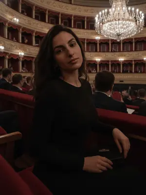 На фоне величественного Большого театра, я уютно расположилась в ложе, окруженной рядами других лож и огромной люстрой из хрусталя. Люди заполняют соседние места, а красные занавески из бархата создают атмосферу изысканности. Камера захватывает момент на уровне моих глаз, создавая впечатление, будто это фотография, сделанная на смартфоне: естественное освещение, нежная пикселизация, минимум ретуши, настоящий момент, неподдельный и живой. Мой взгляд направлен точно в объектив, с легкой улыбкой на губах. Голова слегка наклонена в сторону, придавая образу небрежную изящность. Волосы сверкают на свету, макияж природный, а губы блестят нежным глянцем. Одна рука ласково прикосается к краю ложа, а в другой у меня маленькая черная сумочка. Весь снимок окутан приглушенным светом, создающим атмосферу загадочности и элегантности. 