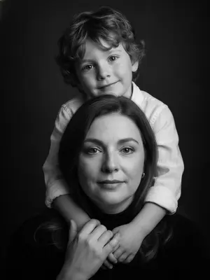 Изображение показывает родителя и ребенка, которые участвуют в семейной фотосессии. Фото выполнено в студии и имеет вертикальную композицию, с крупным планом лиц. Родитель, находящийся внизу кадра, направляет свой взгляд прямо в объектив, лицо полностью обращено к нам. Ребенок, находящийся сверху, повернул голову в сторону, его щека лежит на плече родителя, и он смотрит в камеру с нежной улыбкой. Рука ребенка обнимает шею родителя, видна в кадре, а родитель, в свою очередь, держит эту руку своей ладонью.

Одежда ребенка состоит из белой рубашки из льна, в то время как родитель одет в черную водолазку. Фон студии выполнен в темно-серых почти черных тонах, что создает контраст с яркими лицами на переднем плане. Освещение мягкое и боковое, оно придает изображению объем и глубину.

Фотография выполнена в черно-белой гамме с выразительными контрастами. Атмосфера, царящая на снимке, пропитана нежностью, интимностью и спокойствием. 