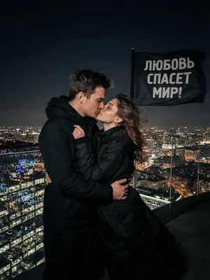 Изображение: Молодая пара, одетая в черное, стоит на вершине высокой башни, обнявшись и целуясь. Они стоят против ветра, который развевает их волосы и одежду. Рядом с ними развевается черный флаг с надписью «ЛЮБОВЬ СПАСЕТ МИР!». Вокруг них виднеется городской пейзаж с высокими зданиями и огнями, отражающимися в стеклянных окнах. Мужчина и женщина выглядят решительными и страстными, их лица освещены лунным светом, подчеркивающим контуры их фигур и выражение лиц. 