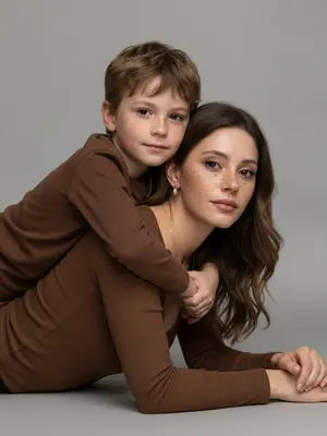 Create a studio portrait capturing the essence of a mother and child, maintaining their original facial features and expressions with precision. The scene is set in a close-up perspective at eye level. The duo is depicted lying on their stomachs, facing sideways to the camera. The child lovingly embraces the mother from behind, their arms wrapped tenderly around her shoulders. Both subjects gaze directly at the viewer, establishing a heartfelt connection. 

The mother exudes a natural beauty with a flawless, matte complexion complemented by subtle makeup - soft pink lips and a hint of mascara. She is dressed in a snug brown long-sleeved top, accentuated by a graceful silver chain and pendant draping elegantly along her collarbone. 

The child mirrors the mother's attire with their own brown long-sleeved top. They wear a delicate, thin chain necklace that harmonizes with the mother's jewelry, adding a touch of visual unity to the composition. 

The image is formatted in 9:16 ratio and rendered in stunning 8K quality. 