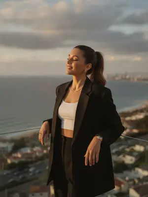 A woman stands gracefully on a balcony overlooking the ocean, bathed in the warm glow of the setting sun. Her body is slightly angled, one arm casually resting on the glass railing as she basks in the sunlight with her eyes closed, exuding an air of effortless elegance.

She is dressed in an oversized black blazer paired with a fitted white crop top and elegant black trousers, adorned with minimal yet luxurious jewelry and a sleek watch that adds a touch of old-world sophistication to her ensemble.

Her makeup is a soft glam look, enhancing her bronzed skin with a natural contour, defined brows, and long, fluttering lashes. The warm sunlight accentuates the realistic texture of her skin, casting soft highlights that add to her radiant glow.

The lighting captures the golden hour ambiance of the sunset, creating a cinematic glow that bathes her in a warm, ethereal light. The soft shadows and highlights play delicately on her hair and skin, enveloping her in an aura of luxury and tranquility.

In the background, the ocean stretches out beneath a soft, cloudy sky, with the city's coastline and roads below blurred into a dreamy backdrop that adds depth to the scene. The overall atmosphere is one of relaxation and sophistication, capturing a moment of pure serenity and beauty.

The image is captured with an iPhone, giving it a realistic and candid feel with a 50mm lens look that focuses on the woman while softly blurring the background. The emphasis is on creating an ultra-realistic portrait that exudes a luxury influencer vibe, showcasing natural body proportions and skin texture with a film-like quality that avoids overprocessed artificial enhancements. 