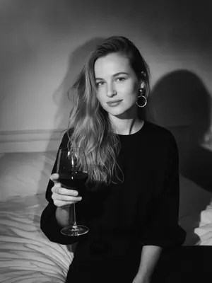 Capture a black-and-white portrait in the Y2K style, taken at night with a direct flash. The subject, a woman, sits gracefully on a tousled white bed, viewed from a slightly raised perspective. She is adorned in a chic black top featuring billowy sleeves and striking circular earrings. One hand delicately cradles a sophisticated glass of deep red wine. Her body subtly sways, imparting a natural blur to her face and shoulders, as if frozen mid-movement. Her gaze is tender, meeting the camera with a soft, intimate smile.

Flowing locks cascade in loose waves, with stray strands gently brushing her face, tousled by her motion. The lighting is stark, casting sharp shadows against the night backdrop, creating a high-contrast ambiance with intense highlights and deep shadows, accentuated by visible film grain. Her skin appears authentic and textured, untouched by digital smoothing, enhanced by the flash's glare.

The overall atmosphere exudes a high-fashion editorial vibe, blending Parisian elegance with a sense of old-world allure. The scene evokes a melancholic nighttime sentiment, capturing a spontaneous and cinematic essence. Shot in a raw 35mm film style, the image boasts pronounced motion blur, adding a touch of lived-in realism to the captivating composition. 