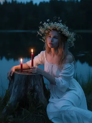 In the tranquil twilight by a serene lake, a woman is seated next to an ancient tree stump. Her posture exudes a dreamy relaxation, leaning slightly forward, one arm casually draped over her knee, the other hand delicately cradling a slender crimson candle beside another flickering flame on the stump. She wears a vintage off-shoulder peasant dress in white, adorned with intricate lace accents and gentle fabric drapes. Her hair is styled with a natural softness, featuring loose strands framing her face.

Adorning her head is a lavish crown of wildflowers, crafted from chamomile blooms, meadow grass, and delicate white blossoms. The background is enveloped by a dark forest and the placid waters of the lake, while the warm glow of the candles casts a gentle light on her face and hands, creating deep blue shadows in the mystical ambiance of a summer night.

Captured with the night mode of an iPhone, the image boasts a realistic grain in low light, with adjusted exposure to maintain sharpness and clarity, devoid of flash, blur, or bokeh effects. 