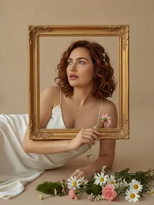 Женщина в белом шелковом платье, украшенном тонкими бретелями, изящно устроилась на светлой поверхности, опираясь на одну руку. Ее черты лица не подверглись изменениям, а макияж был выполнен естественно: глянцевые губы ягодного оттенка, легкий румянец и сияющая влажная кожа. В ее волосах украшение из маленьких белых цветов, а в руке она держит нежный розовый цветок. Ее взгляд направлен вверх и в сторону, губы слегка раскрыты, лицо выражает мечтательность.

Перед женщиной расположена большая декоративная золотая рама, через которую просматривается ее лицо и верх тела, создавая впечатление "картины внутри кадра". На переднем плане размещена композиция из живых цветов: белые ромашки, розовые гвоздики, мелкие полевые цветы и зеленый мох. Фон состоит из бежевой студийной стены, лишенной деталей, создавая минималистичный вид. Теплое освещение, идущее сбоку, придает коже золотистый оттенок и легкие тени. Фотография обладает кинематографической мягкостью, неглубокой глубиной резкости, легким зерном и естественной текстурой кожи. Цветовая коррекция теплая, придающая золотистый оттенок. Композиция наполнена художественным, романтичным и слегка винтажным стилем, с низким контрастом и мягким фокусом, создавая этерическую эстетику. 