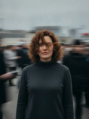 На фотографии, созданной с эффектом размытого движения при помощи Leica SL3 и объектива 50mm Summilux на диафрагме f/2.8, запечатлен момент с фронтального ракурса на уровне груди. Ровный, но пасмурный свет дня действует как нежный диффузор, позволяя создавать плавные следы движения без резких контрастов. Палитра цветов включает в себя приглушенные оттенки бетона и нейтральные тонки кожи, вдохновленные творчеством студии A24.

Человек на снимке: волосы застыли в воздухе перед лицом, кожа выглядит естественной, глаза пронзительно смотрят прямо в объектив, сохраняя спокойствие в окружении хаоса. Выражение лица покойное, сосредоточенное. Одежда подобрана в минималистичном стиле без ярких деталей.

Вокруг множество прохожих, мчащихся в разные стороны, их силуэты растянуты в плавные горизонтальные и диагональные размытия. Все это создает ощущение тихого протеста сквозь неподвижность. Атмосфера на снимке - редакционное спокойствие в противовес хаосу.

Постобработка изображения включает в себя чистую передачу движения, тонкое зерно, отсутствие глитчей, искажений лица и деформаций анатомии. Формат изображения - ar 9:16, сохранен в формате raw. 