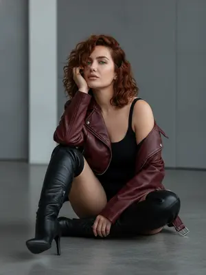 Женщина, выглядящая уверенно, сидит на полу студии. Она одета в свободную темно-красную кожаную куртку, которая спадает с одного плеча, открывая плечо. Под курткой виден черный топ, а также высокие сапоги на шпильке. На ней короткие шорты, но их не видно из-за позы. Фон студии минималистичен, в серых тонах, освещение мягкое. Макияж натуральный и гламурный, с едва заметными контурами. Поза женщины непринужденно-шикарная, соответствующая современной эстетике высокой моды. Стиль фотографии напоминает редакционные съемки Vogue, с ультрареалистичным изображением в 8K качестве. Тон фотографии кинематографичен, композиция сбалансирована. Общее настроение излучает утонченность и минимализм. Голова женщины опирается на руку, сохраняя ее черты лица. 