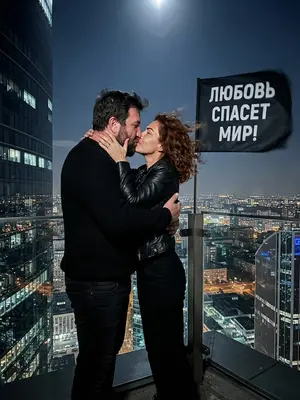 Изображение: Молодая пара, одетая в черное, стоит на вершине высокой башни, обнявшись и целуясь. Они стоят против ветра, который развевает их волосы и одежду. Рядом с ними развевается черный флаг с надписью «ЛЮБОВЬ СПАСЕТ МИР!». Вокруг них виднеется городской пейзаж с высокими зданиями и огнями, отражающимися в стеклянных окнах. Мужчина и женщина выглядят решительными и страстными, их лица освещены лунным светом, подчеркивающим контуры их фигур и выражение лиц. 