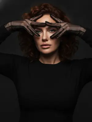 На фотографии тесным кадром запечатлена очаровательная женщина на темно-сером фоне, одетая в прозрачные черные перчатки и черное платье. Она элегантно позирует, обрамляя свое лицо руками, создавая игривые тени вокруг глаз. Ее яркий глазной макияж с аккуратной подводкой, безупречная кожа, кинематографическое освещение и высокомодный стиль добавляют атмосферу загадочности и соблазнительности. 