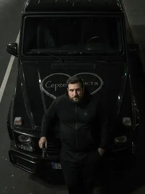 На заднем плане мрачное ночное окружение городской улицы, смутные очертания зданий и фонарей. В центре кадра человек, одетый в черный спортивный костюм без излишних деталей, стоит у автомобиля, наклонившись на капот. В руке у него легко покачиваются ключи от машины. На капоте, занимающем всю площадь кадра, выгравировано грубое сердце с таинственным текстом внутри. Отражение жесткого прямого света создает резкие тени и подчеркивает микроскопические дефекты краски, словно пыль и царапины. Съемка выполнена с верхнего ракурса, придающего кинематографическую атмосферу. Реалистичные текстуры кожи и ткани костюма подчеркиваются объективом 35 мм с диафрагмой f/2.8, создавая грубую уличную эстетику. 