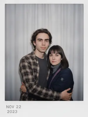 Сделайте снимок с помощью фотоаппарата Polaroid. Фотография должна выглядеть как обычное фото, без четкого объекта или реквизита. На фото должен быть постоянный источник света, такой как вспышка из темной комнаты, равномерно распределенная по всей фотографии. Не меняйте лица. Замените фон за спинами двух человек на белую занавеску. Люди на фотографии обнимаются. 