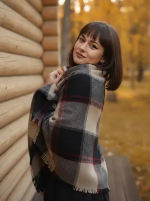На улице, в осенний день, запечатленный в теплых оттенках золота и оранжевого, создается атмосфера уюта и романтики, пронизывающая снимок. На заднем плане виднеется стена из светлых деревянных бревен, за которой раскинулись деревья с яркой желто-красной листвой, придающей изображению эффект размытия. Модель, стоя полубоком к объективу, слегка поворачивает корпус, а ее голова наклонена в сторону, с мягкой улыбкой, направленной на зрителя. Она завернута в объемный, теплый клетчатый плед, в котором гармонично сочетаются черные, серые, бежевые и бордовые оттенки. Края пледа она держит руками у подбородка, скрывая нижнюю часть лица, а под ним угадывается темная одежда - возможно, юбка или брюки. Мягкое, естественное освещение акцентирует внимание на текстуре как пледа, так и деревянных бревен, создавая атмосферу спокойствия и меланхолии. Модель выглядит как настоящая профессиональная фотомодель. 