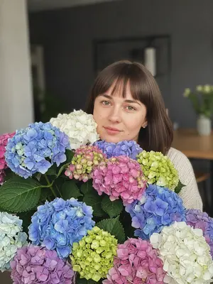 Женщина на портрете окружена огромным букетом цветов, частично скрывающим ее тело, но оставляющим лицо видимым. Камера направлена точно на модель, создавая ощущение личного контакта. Поза естественная: женщина наклонена к цветам, подбородок опирается на верхний угол букета. Ее взгляд прямой, мягкий, с намеком на улыбку, выражая спокойствие и удовлетворение. Букет состоит из более 30 крупных цветов, некоторые раскрыты, другие - в бутонах, создавая динамику. Зеленые стебли и листья свежие, с прожилками и естественным блеском. Фон размытый, показывая элементы интерьера, такие как темные стены и светлая столешница. Естественный свет падает слева, создавая мягкие тени и блики на лице и цветах. Обработка с использованием фильтра «soft floral natural», добавление микрошума для эффекта пленки, усиление контраста для выделения деталей. В целом, эмоциональный тон портрета - радость, благодарность, женственность. Композиция соблюдает правило третей, лицо модели смещено вправо, букет занимает левую и нижнюю часть кадра. Стиль фотографии - современная эстетика Instagram, с акцентом на естественность и эмоции. 
