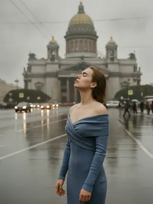 Фотография на улице Санкт-Петербурга после дождя, передающая атмосферу города. На снимке запечатлен человек в стильной зоне. Он стоит у фонарного столба, обрамленного сырым камнем, который отражает отблески влажного асфальта. Человек одет в черное пальто и широкополую шляпу, ладонью касается края головного убора. Его взгляд устремлен вдаль, словно он ожидает кого-то или что-то. Волосы чуть взмокли от оставшихся после дождя капель. На лице бледный свет от фонаря, создающий загадочную атмосферу. Вокруг витает запах свежести, а на фоне зданий виднеется туман, придающий картины загадочности и некой мистической притягательности. 