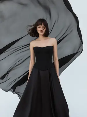 На студийном снимке fashion-портрета ультрареалистичного стиля, крупный план модели от груди до бедер. Модель стоит на белой циклораме, окруженной чистотой. За ней расправлена ткань из черного шифона, плавно колышущаяся под воздействием ветра. Этот элемент создает драматический фон с графичными контурами, ярко выделяясь на фоне белого пространства студии. 