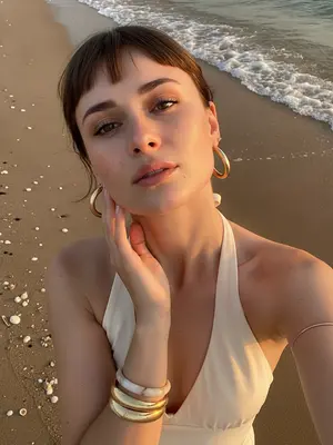 A woman stands gracefully on the sandy beach by the water, capturing a lifestyle beauty moment with her smartphone. The camera looks down on her, giving a high perspective typical of a handheld selfie. She tilts her head slightly upwards, gazing at the camera, while her hand delicately brushes the side of her face, fingers tracing the curve of her cheek and jawline. Her shoulders are relaxed, creating an elegant pose in the frame that captures her from the chest up.

Dressed in a cream halter neck swimsuit or summer top, her open shoulders and exposed collarbones exude a sense of summer chic. Adorning her look are large gold hoop earrings, a thick sculptural gold bracelet, a light marble-style bangle, and a delicate metallic arm bracelet above her elbow. Her makeup is soft and glamorous, perfect for a beach setting: her skin glows with a warm sun-kissed radiance, peach blush accentuates her cheekbones, clean sculpted brows frame her eyes, subtle winged eyeliner enhances her gaze, long lashes flutter, and her lips shimmer with satin finish.

In the background, the wet beach sand offers natural textures, with a thin line of sea foam marking the approach of a small wave behind her. Small shells are scattered on the sand, adding to the coastal atmosphere. The lighting captures the magic of a sunset or golden hour, casting warm highlights on her skin and creating soft shadows that play around her facial features. The shallow depth of field adds to the candid summer vacation mood, blending a realistic DSLR smartphone hybrid aesthetic seamlessly. 