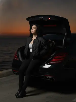 На краю морского пирса стоит черный Mercedes S-Class W222 AMG без номеров, блики луны играют на его кузове. В кадре виден только задний бампер и открытый багажник, из которого торчит часть модели. Девушка сидит на краю багажника, касаясь ладонью металлической детали, ноги покоятся на воздухе, обувь теряется в тени. Руки спрятаны в карманах кожаной куртки, голова слегка откинута назад. Ее взгляд устремлен вдаль, в сторону темно-оранжевого заката над горизонтом. Атмосфера пронизана тишиной позднего вечера, когда ночь уже на пороге. Освещение тусклое, с отсветом вспышки фотоаппарата, создающей реалистичные тени. Снимок сделан на Canon G7X, захватывая естественную позу в живом уличном кадре, словно картина ночного города. 