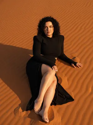 In the soft glow of the golden hour, a woman reclines gracefully on the undulating surface of a sun-kissed orange sand dune. The full view captures her from head to toe, with both feet elegantly displayed, the scene captured from a low angle. Her body is positioned diagonally across the dune, one arm supporting her as the other hand casually rests by her thigh. With one leg extended and the other slightly bent, the high slit of her skirt adds a touch of allure to the ensemble.

Gazing directly at the camera, her expression exudes a sense of calm confidence. She is adorned in a striking black hooded ensemble, featuring structured shoulders, form-fitting long sleeves with thumbholes, a waist cutout, and a floor-length skirt with a daring slit. The makeup is kept minimal, creating a dewy look that enhances her natural beauty. Her clean white manicure and realistic skin texture, complete with visible pores, add to the authenticity of the image.

The warm, golden light bathes the scene, casting gentle shadows and highlighting the intricate textures of the sand and fabric. Shot with a Canon EOS R and a 24-70mm lens, the focus is sharp, capturing every detail with a hyper-realistic quality. The overall atmosphere is cinematic yet natural, evoking a sense of editorial elegance in the desert setting. 